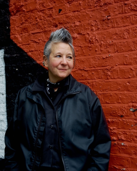 Spike Spondike portrait, wearing a black jacket over a black button-down with a black bandanna tied around her neck, in front of a rust-red wall.